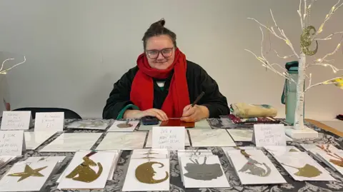 Megan Fatharly A brunette lady with a red scarf on sat behind a desk full of gold a silver artwork in the shapes of stars, dogs, a moon and others.