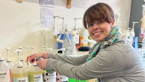 Stephanie Reed A woman in a grey top and multicoloured scarf at a refill store smiles as she turns her head towards the camera. 
