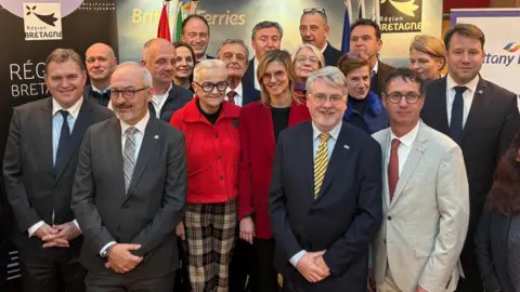 A group photo of 18 men and women smiling at the camera. Behind them are banners reading REGION BRETAGNE and BRITTANY ferries.