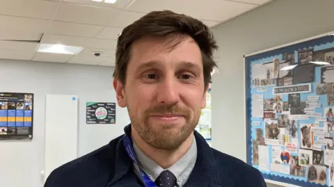 A photo of a man in a classroom with pictures on a noticeboard in the background