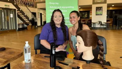 Two women with long dark hair sit at a wooden table with a mannequin head in front of them, which has its hair tied in a braid. They are in a large hall with a wooden floor and a green sign behind them.