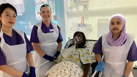 BBC Three student nurses in scrubs smiles next to a manikin doll wearing an oxygen mask with brown hair leans back against a pillow