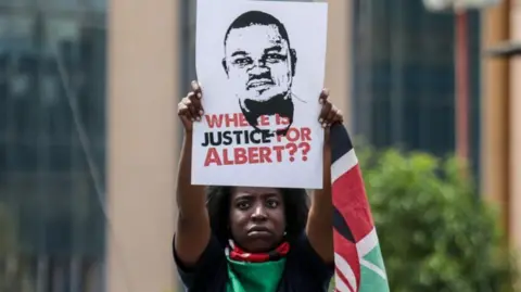 A protester wrapped in a Kenyan flag holds a portrait of Albert Ojwang with the words 'Where is justice for Albert??'