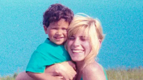 A young child with curly hair is being held closely by an adult with light-coloured hair; both are positioned outdoors against a bright blue sky, with grassy landscape visible in the background.