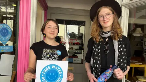 BBC A girl stands on the left holding a sign saying 'free water'. She's wearing a black printed t-shirt and has short hair. On the right, a taller boy is wearing a black hat, scarf and shirt, holding a purple water bottle. They're stood in a shop doorway.