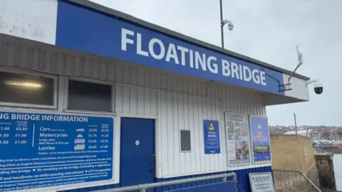 Image shows the boarding area for the Cowes chain ferry challenge. There is a sign that says 'Floating Bridge' and is blue and white in colour. Underneath there is a sign that advertises the cost for cars, motorcycles and lorries. 