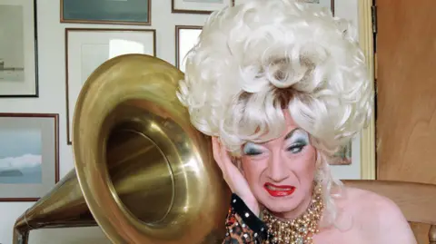 Paul O'Grady as his alter ego Lily Savage in a blonde wig next to a grammarphone 