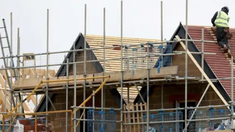 A house under construction surrounded by metal scaffolding. A builder wearing a high-visibility vest, safety helmet and work boots is on the sloping roof at the top right of the image. Part of the roof is covered with tiles, the other is covered in timber battens.