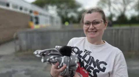 A woman wearing a white 'The Rolling Stones' branded top is standing outside a community centre holding a kitten. The kitten is wrapped in a blanket. The woman is smiling at the camera. 