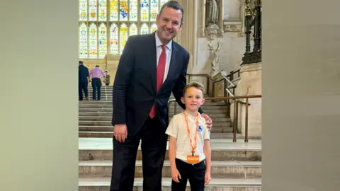 Teddy standing on a stone staircase with a stained glass window behind. The six-year-old boy is wearing a yellow shirt with an orange lanyard around his neck. He is standing with MP Paul Holmes who has his arm around Teddy's shoulder. He is wearing a dark suit with a red tie.