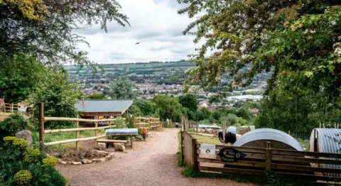 A path between some pens with wooden fences at Bath City Farm. Houses and green hills can be seen in the distance. 