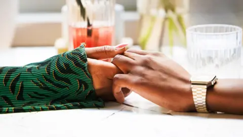 Getty Images Two hands holding across a table, one has a gold watch on, the other has a green long sleeved top. There are some drinks visible in the background.
