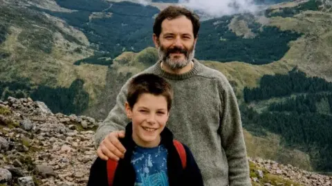 A young boy standing on top of a hill with a blue t-shirt on. The straps of a red backpack are visible over his shoulders. A man with a thick beard has his arm round the boy. They are both smiling.