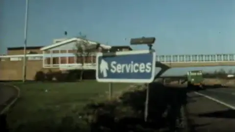 A 1970's TV screen grab showing a single-storey brick building next to a motorway with a footbridge linking sites on the two carriageways and a blue sign directing drivers towards "Services".