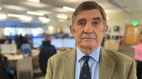 Philip Wilkinson looks into the camera, wearing a tweed jacket, blue shirt and tie, Blurred behind him is a busy office.