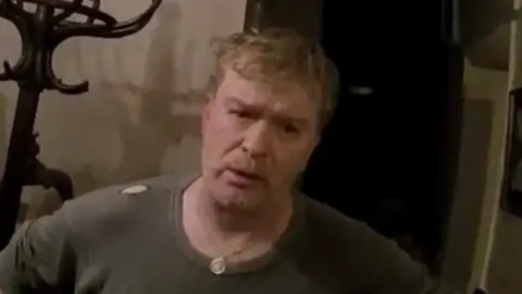 gmp Traynor listens to a police officer with his hands on his hips. He wears a dark top with a chain and stands in a room with a coat stand behind him.