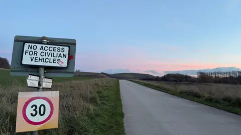 A concrete road reaching into the distance, where there are two hills rising out of the landscape at dusk. To the left in the foreground, a road sign with a 30 mile limit, but also a sign saying 'No access for civilian vehicles.