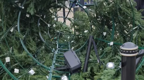 Jade Aubin The inside of a cone-shaped artificial Christmas tree, lying on its side on a pavement. It looks like a metal-framed structure, with a white sandbag under the front section. Silver baubles are attached to the green branches. It has a lightweight webbing cordon around it and there is a black and gold bollard on the right