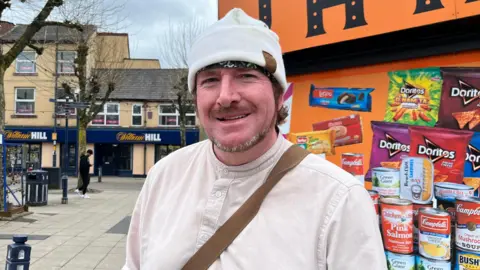 Jonny Humphries/BBC Pau Bruffell, 58, smiles at the camera in front of a shop sign advertising different products. He has a short light brown beard and is wearing a white woollen hat and a white linen shirt. 