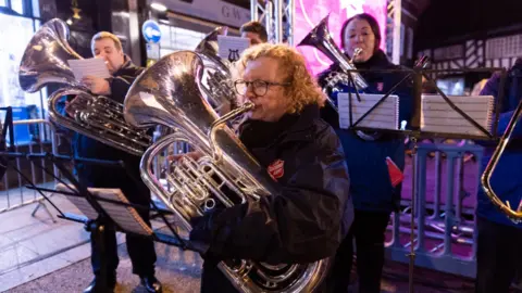 Andy Burton/Worcester City Council A woman with blonde curly hair plays in a brass band - she's playing the tuba. She's also wearing a black rain coat with the Salvation Army emblem on her chest. 