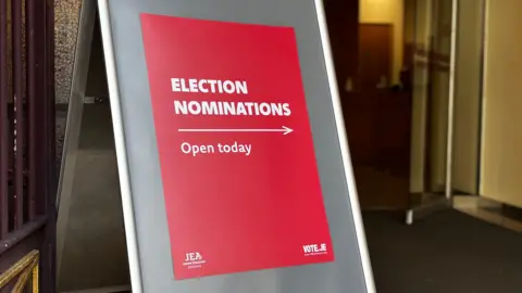A red sign, with white writing saying 'election nominations open today' alongside an arrow pointing to the right. It placed outside a building.