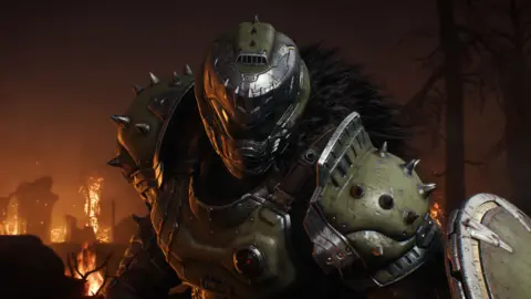 ID Software Artwork showing the Doom Slayer, the main character from the Doom series. He leans towards the viewer, his hulking frame covered by battle-marked green armour, and his face obscured by his trademark helmet and visor. Fires burn in the background.