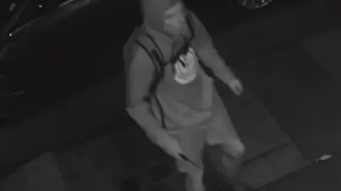 Met Police Black and white image of the suspect wearing shorts, a hoodie with a round logo, backpack and trainers