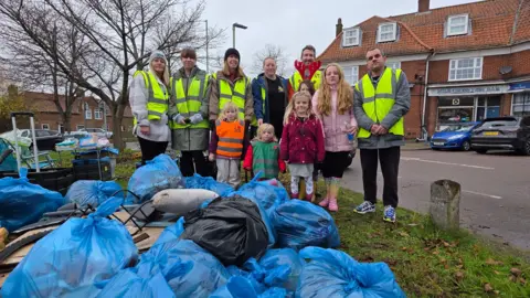 Paul Moseley/BBC Four women and two men, all wearing high-vis jackets, with four children posing for a photo. In front of them are a large number of blue bin bags.