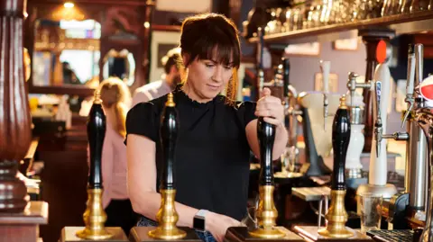 Getty Images Woman with dark brown hair wearing a black short-sleeve top pours pint or beer with pump behind a bar in a pub