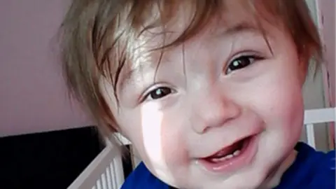 Ethan with brown hair which is sweeping over his forehead. He is wearing a blue top and is in a white cot as he smiles at the camera, his front baby teeth showing.