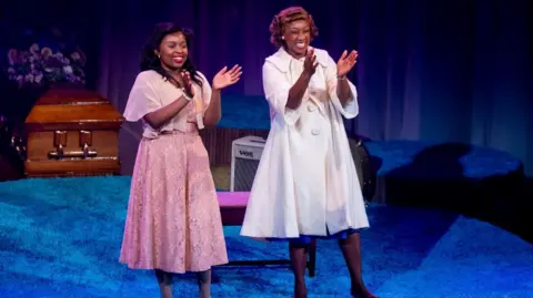 Jonathan Hipkiss Two women standing on a stage smiling. The woman on the left is wearing a pink lace dress and cape. The one on the right is wearing a fifties style flared white coat and is clapping. The stage is covered in a royal blue carpet and there is a wooden casket behind the women.
