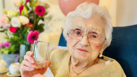 Maisie Dalziel is seated in a blue chair and holding her drink up to the camera in a cheers pose. She has short white hair and is wearing glasses. She has a gold coloured top on and there is a large bunch of flowers behind her.