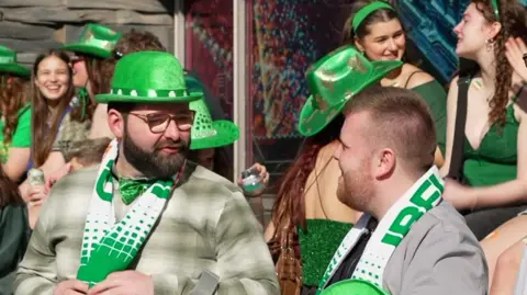 A bearded man with a green pork pie hat and green and white scar talks with his friend who has removed his green hat but also has a similar scarf. Girls in green stetsons are in the background.