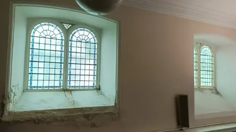 Sandra Pratt Interior of the church building looking at a wall and two windows with stained glass. The walls surrounding the windows are showing signs of damage and damp. The walls are light pink in colour.