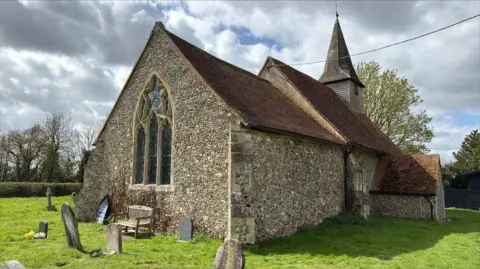 Richard Knights/BBC A view of the rear of a medieval church, showing a large leaded window, and a small steeple at the opposite end. The roof is tiled red. It is surrounded by short grass, with a bench and old gravestones in the foreground. 