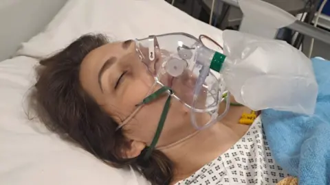 Brunette woman lying down in hospital bed with oxygen mask over her face and eyes closed.