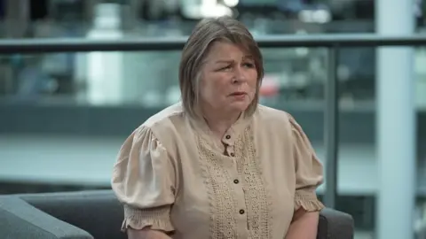 A woman in a cream top and shoulder length hair is looking away from the camera. She is sat on a grey chair and there is a divider made of glass in the background. 