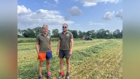 Two men stand in a field. It is a sunny day. They are wearing shorts. There is a line of freshly cut hay behind them. 