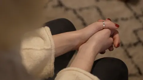 A changeable  from above, showing a woman's hands clasped together. Wearing reddish  nail varnish and wearing a metallic  ring.