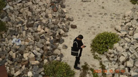 Lily Radwan A man standing outdoors surrounded by rubble and piles of rocks, with a few wild plants and bushes growing around him. He is looking down at his phone, and is carrying a tote bag and wearing all black.