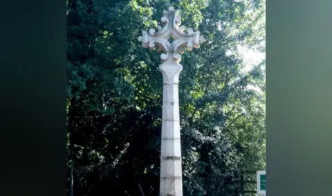 Warwickshire Police A tall, thin pale stone monument stands upright near some trees, and atop it is an ornate cross shape. 