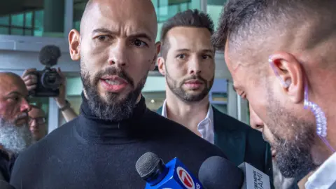 Andrew Tate speaks to a reporter, with his brother Tristan standing behind him