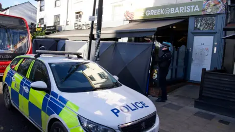 John Fairhall/BBC A police officer stands in front of black screens and a police car at the Barista venue, which has a black canopy and green banner across its entrance. It is based on a busy city street scene.