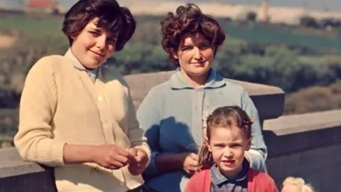 A young woman with short dark hair wearing a cream cardigan leans against a wall smiling at the camera. Another young woman with short dark hair stands next to her wearing a blue cardigan with a six year old girl standing in front of her wearing a blue collared dress with pink cardigan and holding a blonde haired doll.