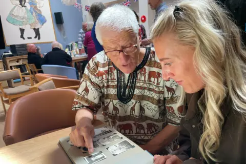 Former Ilford Park resident Charles Jagla and his granddaughter. Jagla is wearing an African style top with safari animals on it. His grandaughter has long blonde hair. They are both looking at old pictures. 