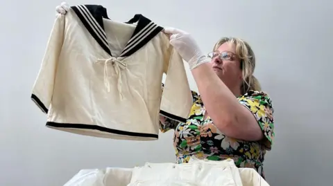 Leeds City Council A woman wearing glasses and a floral dress holds up a children's white sailor suit top. It has navy blue trimmings, a v-neck and a bow tied across the chest.