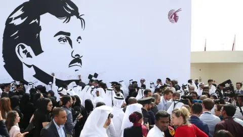 AFP Qatar Airways workers stand in front of a wall bearing a portrait of Qatari Emir Sheikh Tamim bin Hamad Al Thani in support of the country and its leader in Doha. 13 July 2017