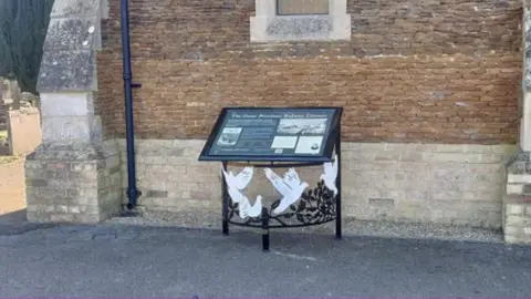 Huntingdon Town Council Black-framed interpretation board alongside stone wall of cemetery chapel