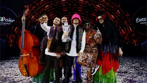 Reuters The six male members of Kalush Orchestra dressed in bright costumes holding the iconic glass microphone Eurovision trophy following their 2022 victory in Turin