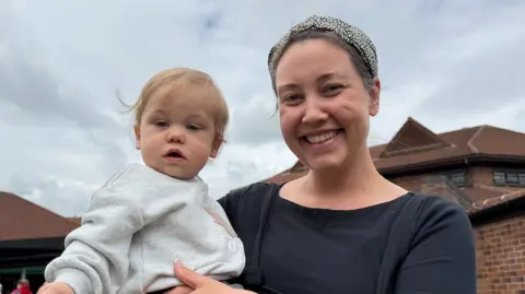 Danielle Moore, wearing a black cardigan and a patterned head scarf, holds her young son, Ozzy, who has blonde hair and is wearing a light grey jumper. 